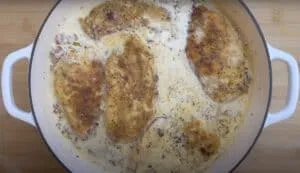 Creamy chicken breast fillets cooking in a white sauce with herbs in a white skillet, perfect for a delicious dinner.